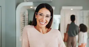 A smiling woman with glasses stands in a room with a blurred background of people and plants