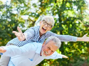 Two older adults, woman sitting on the man's back, are laughing and posing for a photo in a park in Idaho Falls.