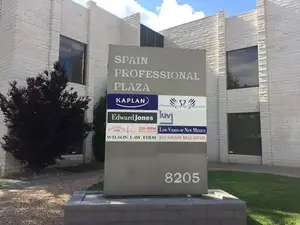 A building with a sign that reads Spain Professional Plaza, it has many glass windows, and a sign board is in front with logos of Kaplan, Edward Jones, Wilson Law Firm, and others, a tree is on the left side, and the number 8205 is at the bottom of the board