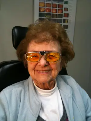 An older woman wearing a pair of sunglasses with a blue sweater and a white turtleneck sitting on a black chair.