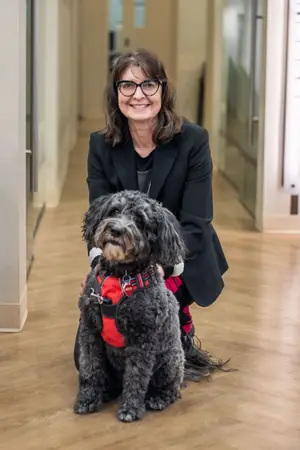 A woman in a black coat and glasses is crouching next to a gray dog with a red harness in a hallway.
