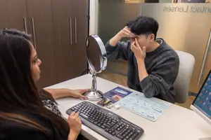 A man is putting in contact lenses with the help of a woman at a desk with a mirror and keyboard.