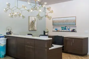 The front desk of a modern office with wooden cabinets, a chandelier, and various items on the wall, including a framed picture of a peacock and a shelf with various items. The desk has a trash can, a flower vase, and a computer monitor. The room has a wooden floor and a white wall.