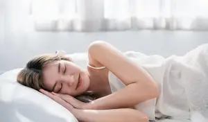 A woman sleeping peacefully on a bed with a serene expression, wearing white clothing, and surrounded by a white room with sunlight filtering through the windows.