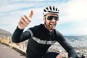 A man in a black cycling outfit and helmet rides a bike and gives a thumbs up on a mountain road with a cityscape in the background.
