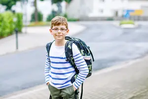 A young boy with glasses and a backpack standing on the sidewalk
