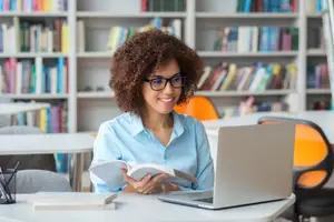 A woman with glasses is reading a book in a library.