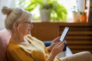 An elderly woman is sitting on a couch, wearing glasses, and looking at her tablet with a smile on her face. Behind her, there is a wooden cabinet with potted plants on top, and the sunlight is shining through the window.