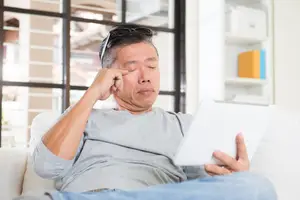 A man is sitting on a couch holding a tablet with one eye closed.