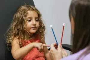 A young girl with curly hair is pointing at a red and blue object, likely a toy, while sitting on a chair with a woman in a purple shirt who is also holding the object.