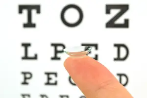 A finger holding a clear contact lens in front of an eye chart.