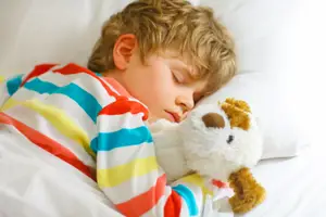 A young child sleeps soundly in bed with a teddy bear, wearing colorful striped pajamas and a peaceful expression.
