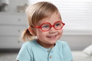 A young girl wearing red-framed glasses and a blue shirt is smiling indoors.