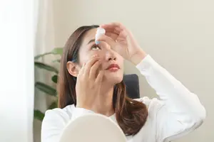 A woman is applying eye drops to her left eye in a well-lit room.
