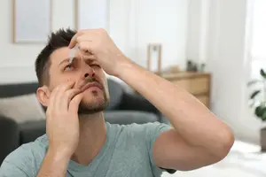 A man uses eye drops while seated indoors, with a couch and plant in the background.