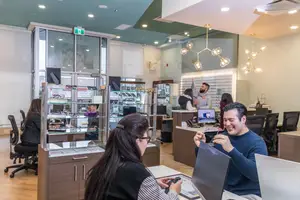 Customers are shopping for glasses at an optometrist office. They are sitting at a desk and trying on frames. Behind them is a glass display case with glasses. In the background, there are other customers and employees.