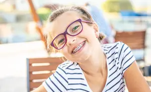 A young girl with braces smiles brightly while wearing glasses and a striped shirt, sitting on a wooden chair outdoors in a sunny setting.
