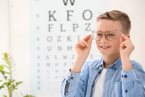A young boy in a blue shirt is adjusting his glasses while smiling at a vision chart in a room.