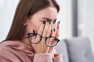 Closeup of a young woman sitting on a couch rubbing her eyes while holding her glasses