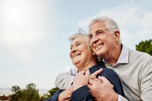 An elderly couple is smiling and hugging each other under a blue sky with clouds