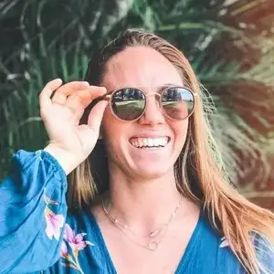 A woman with blonde hair is wearing a blue floral dress and sunglasses. She is smiling and posing for a photo while holding her sunglasses up to her face.