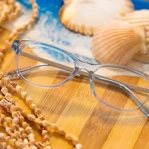 A pair of clear glasses with a silver frame and curved edges is placed on top of a wooden table with a plate of cookies and seashells. A beaded necklace with a seashell pendant is on the left side of the table, and the seashells have a slightly curved shape.