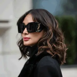 A woman with brown wavy hair wearing a black coat and black sunglasses with a gold YSL logo on the side.