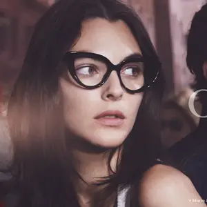 A woman with long black hair is wearing black cat-eye glasses and a white sleeveless top, looking into the distance.