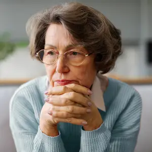An elderly woman wearing glasses and a light blue sweater is sitting on a couch and has her hands clasped together while looking down.