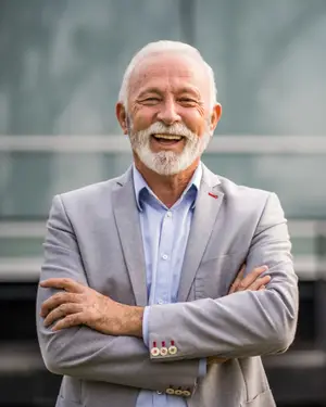 A senior man with a white beard is smiling and standing with his arms crossed, wearing a gray suit and a blue shirt with a red logo on the left side of his chest.