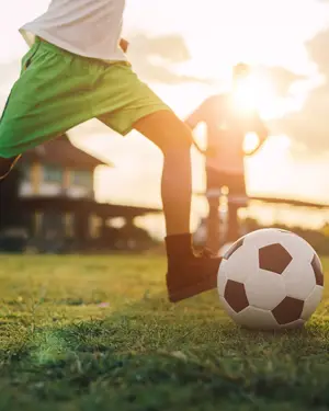 A soccer player in green shorts is kicking a soccer ball on a grassy field, with another person standing behind him, while the sun is shining brightly in the background.