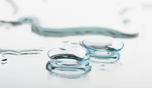 Two transparent contact lenses sit in their solution on a white surface, one with a small air bubble.
