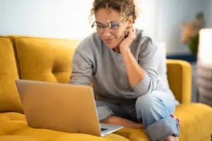 Woman sitting on a couch while talking on the phone and looking at a laptop.