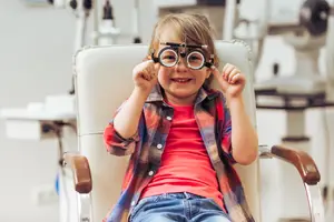 A little girl in a red shirt and jeans with a plaid shirt over it is sitting in a chair wearing an eye test instrument and smiling for the camera.