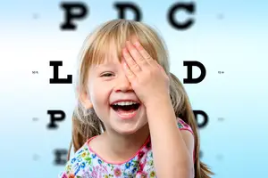 A young girl is smiling and covering her left eye with her hand in front of a blue background with letters on it.