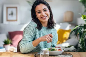A smiling woman with a salad and fork sitting at a wooden table in a home setting
