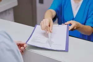 A woman in a blue uniform is filling out a form with a pen while a man is looking at her in a hospital setting.