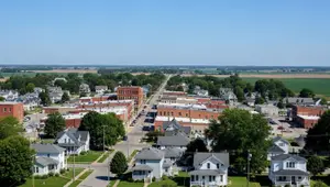 A town with several houses and buildings, and a road in the middle. There are also trees and plants in the area. The sky is clear and blue.