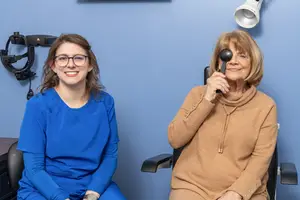 Two women in a medical office, one holding an eye exam tool, smiling at the camera.