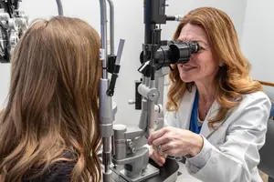 A female optometrist examines a patient's eyes using an ophthalmoscope in a clinic setting.