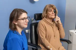 A woman in a brown sweater is having her eye examined by a woman in a blue shirt in a clinic