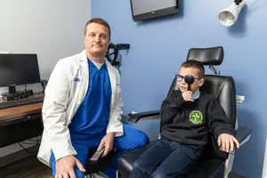 A doctor in a white coat is smiling at a young boy in a black jacket with a logo on it, sitting in a black chair in a blue room with a monitor, keyboard, and television on the wall.