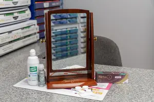 A display of eye care products, including a mirror, Opti-Free solution, Biotrue contact lens solution, and Total 1 contact lenses, set on a counter in a room.