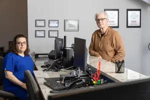 Two people, one sitting and one standing, pose in front of a desk with computers and various office supplies.