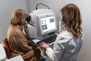 A woman sitting on a chair in front of a monitor and a doctor using a keyboard, they are probably doing an eye checkup