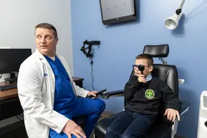 A man in a white coat is sitting in a chair and holding a remote control in his hand while a boy in a black sweater is sitting on a chair in front of him and wearing glasses