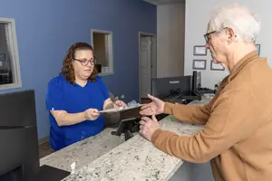 An elderly man in a brown sweater is speaking with a woman wearing a blue shirt and glasses at a reception desk in a medical office.