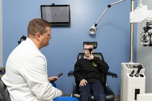 A boy in a black coat is getting his eyes checked by a doctor in a white coat while sitting in an eye examination chair.