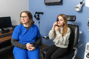 A young girl wearing eye patches is sitting on a chair and smiling while the woman next to her is looking at her. There is a monitor and keyboard on the table, and a lamp is on the wall.