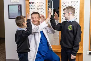 A man in a white coat and blue pants is doing a high five with two boys in black shirts in front of a display of glasses